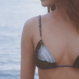 A woman wearing the Dolores Top Classic Swimwear, featuring delicate jewelry straps, stands near the water; her face is partially visible as a calm, blurred ocean forms the background.