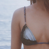 A woman wearing the Dolores Top Classic Swimwear, featuring delicate jewelry straps, stands near the water; her face is partially visible as a calm, blurred ocean forms the background.