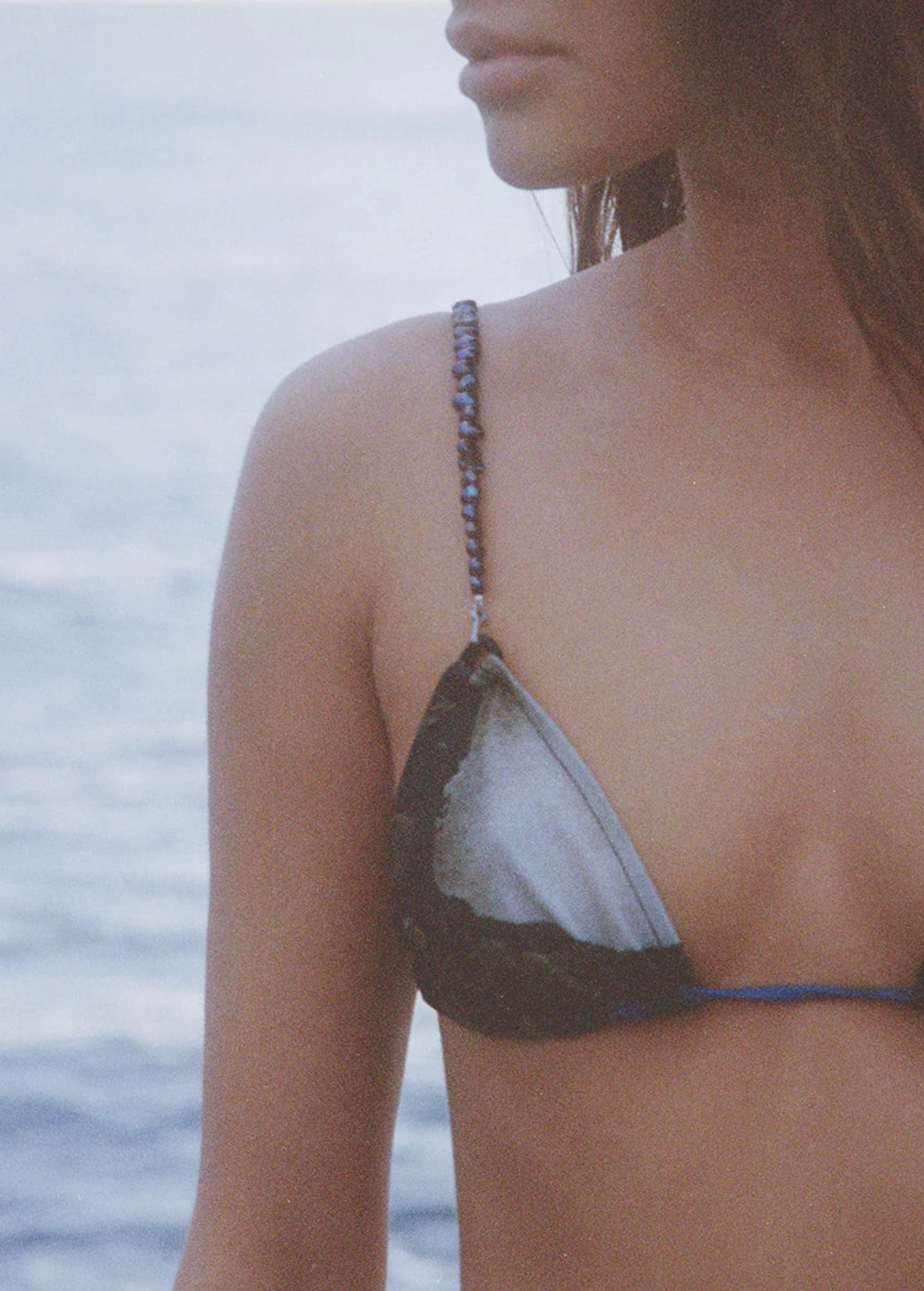 A woman wearing the Dolores Top Classic Swimwear, featuring delicate jewelry straps, stands near the water; her face is partially visible as a calm, blurred ocean forms the background.