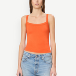 A woman wears a bright orange cotton-modal Duke Tank with a square neckline and blue denim shorts, styled for everyday wear.
