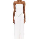 A person stands against a white background wearing The Triangle Top—a strapless piece with a pointed hem—paired with wide-leg white pants. This ensemble reflects the artistry of handmade clothing and an appreciation for sustainable fashion, while transparent glasses complete the effortlessly modern look.