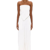 A person stands against a white background wearing The Triangle Top—a strapless piece with a pointed hem—paired with wide-leg white pants. This ensemble reflects the artistry of handmade clothing and an appreciation for sustainable fashion, while transparent glasses complete the effortlessly modern look.
