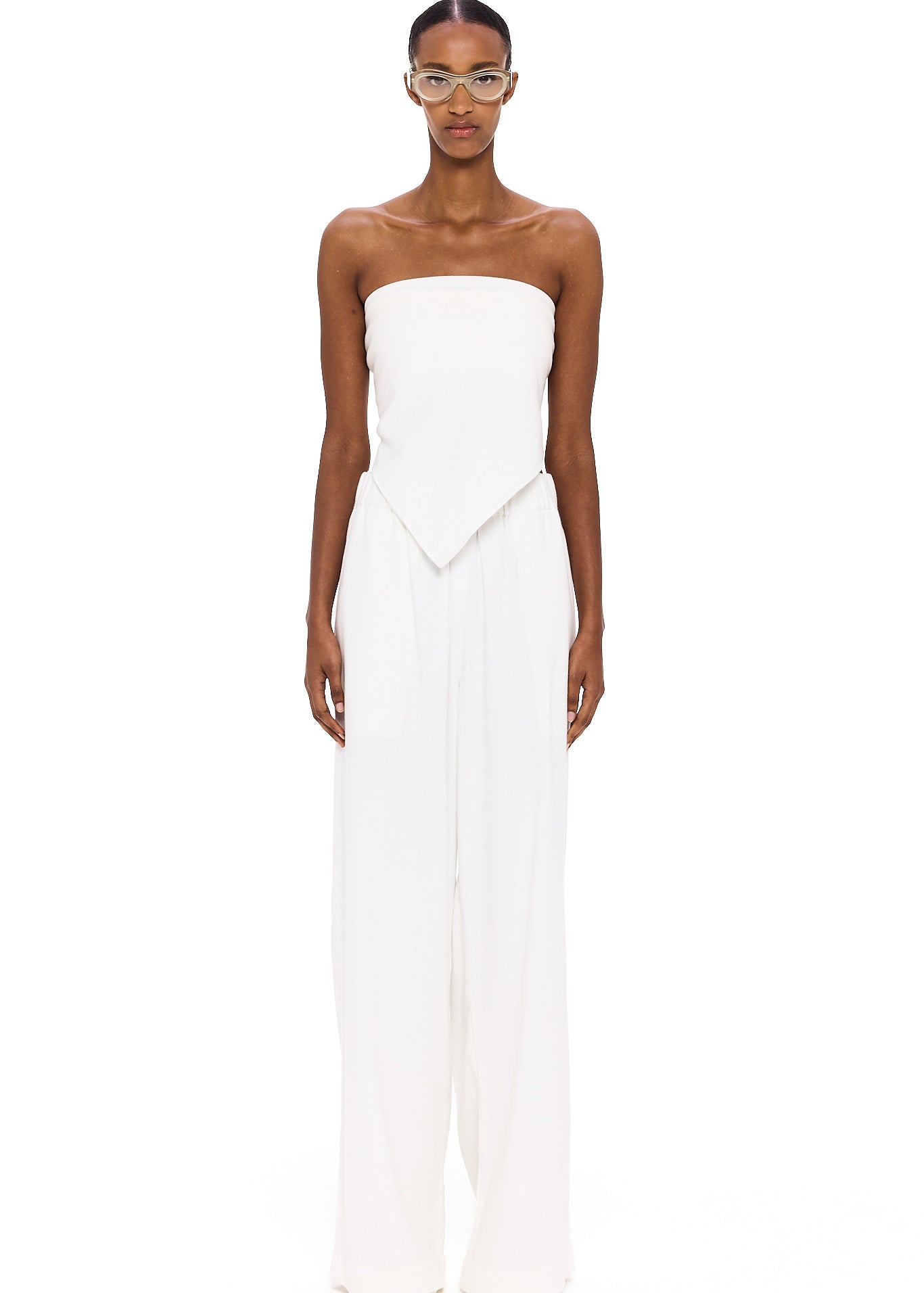 A person stands against a white background wearing The Triangle Top—a strapless piece with a pointed hem—paired with wide-leg white pants. This ensemble reflects the artistry of handmade clothing and an appreciation for sustainable fashion, while transparent glasses complete the effortlessly modern look.