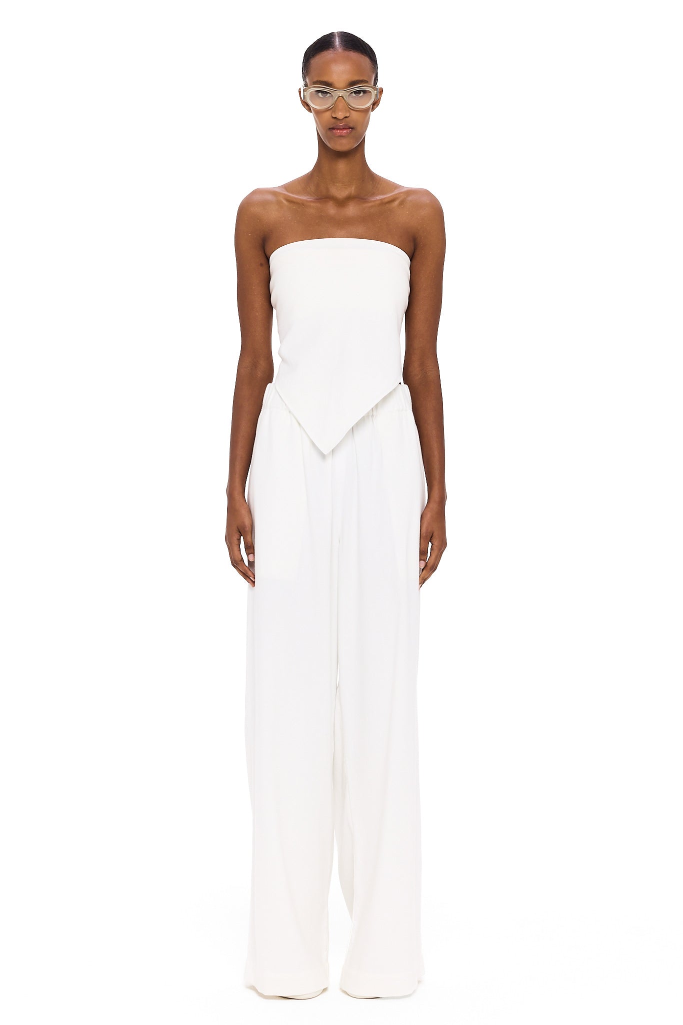 A person stands against a white background wearing The Triangle Top—a strapless piece with a pointed hem—paired with wide-leg white pants. This ensemble reflects the artistry of handmade clothing and an appreciation for sustainable fashion, while transparent glasses complete the effortlessly modern look.
