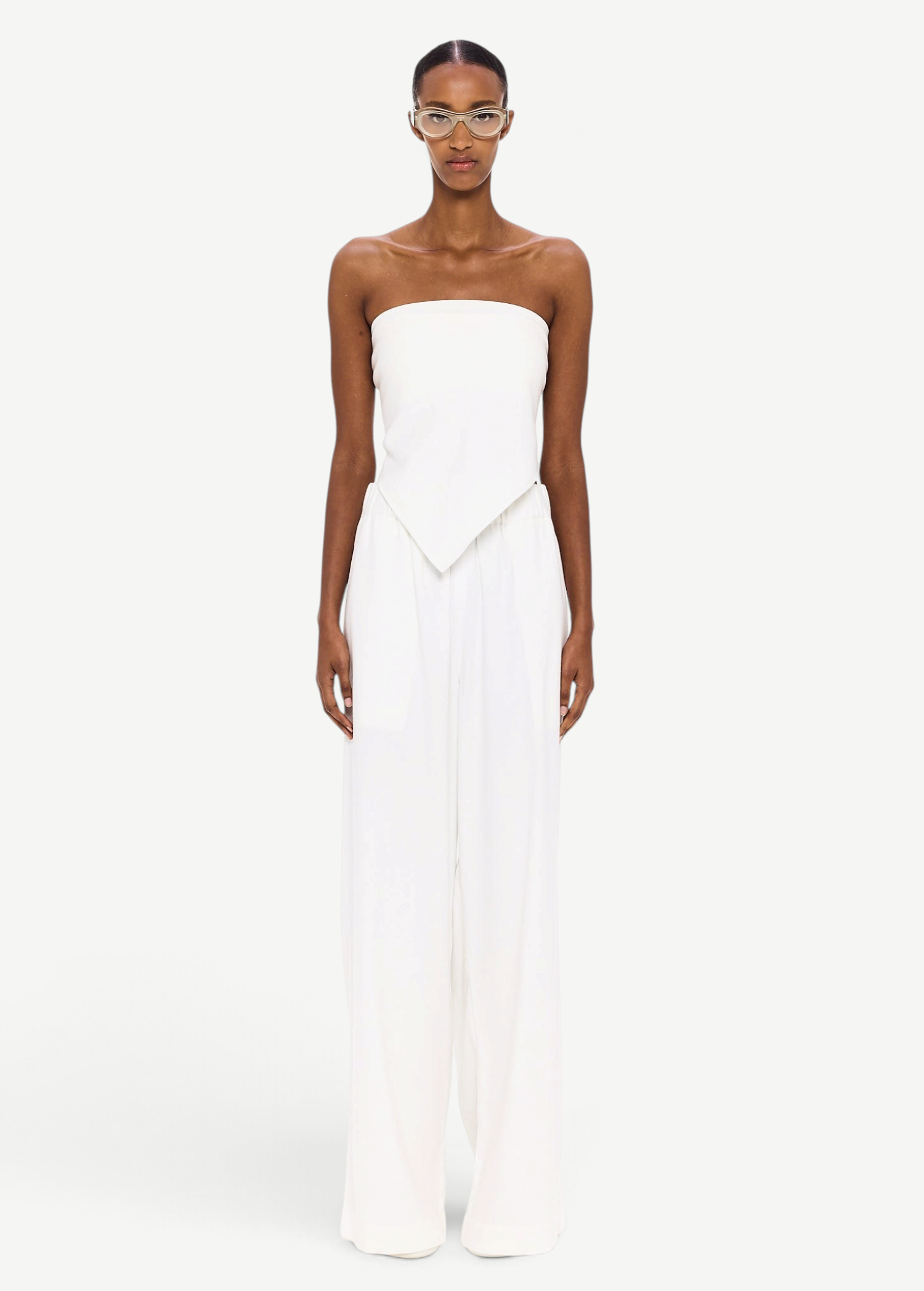 A person stands against a white background wearing The Triangle Top—a strapless piece with a pointed hem—paired with wide-leg white pants. This ensemble reflects the artistry of handmade clothing and an appreciation for sustainable fashion, while transparent glasses complete the effortlessly modern look.
