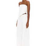 Dressed in The Triangle Top—a strapless white piece featuring an uneven hem—paired with wide-legged white pants, this individual stands against a white background, looking forward while wearing glasses; a modern showcase of sustainable fashion and the artistry of thoughtfully crafted, handmade clothing.