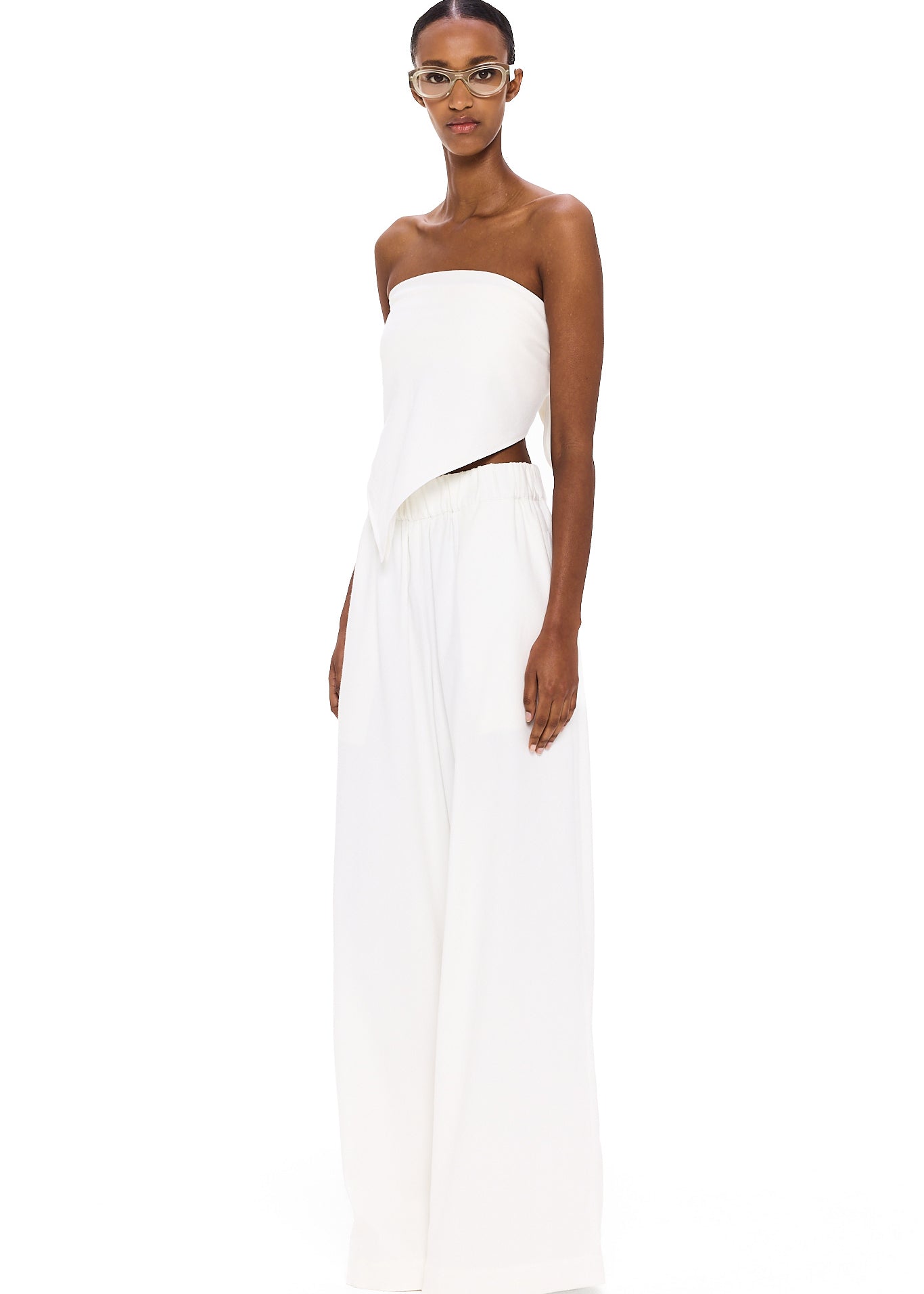 Dressed in The Triangle Top—a strapless white piece featuring an uneven hem—paired with wide-legged white pants, this individual stands against a white background, looking forward while wearing glasses; a modern showcase of sustainable fashion and the artistry of thoughtfully crafted, handmade clothing.