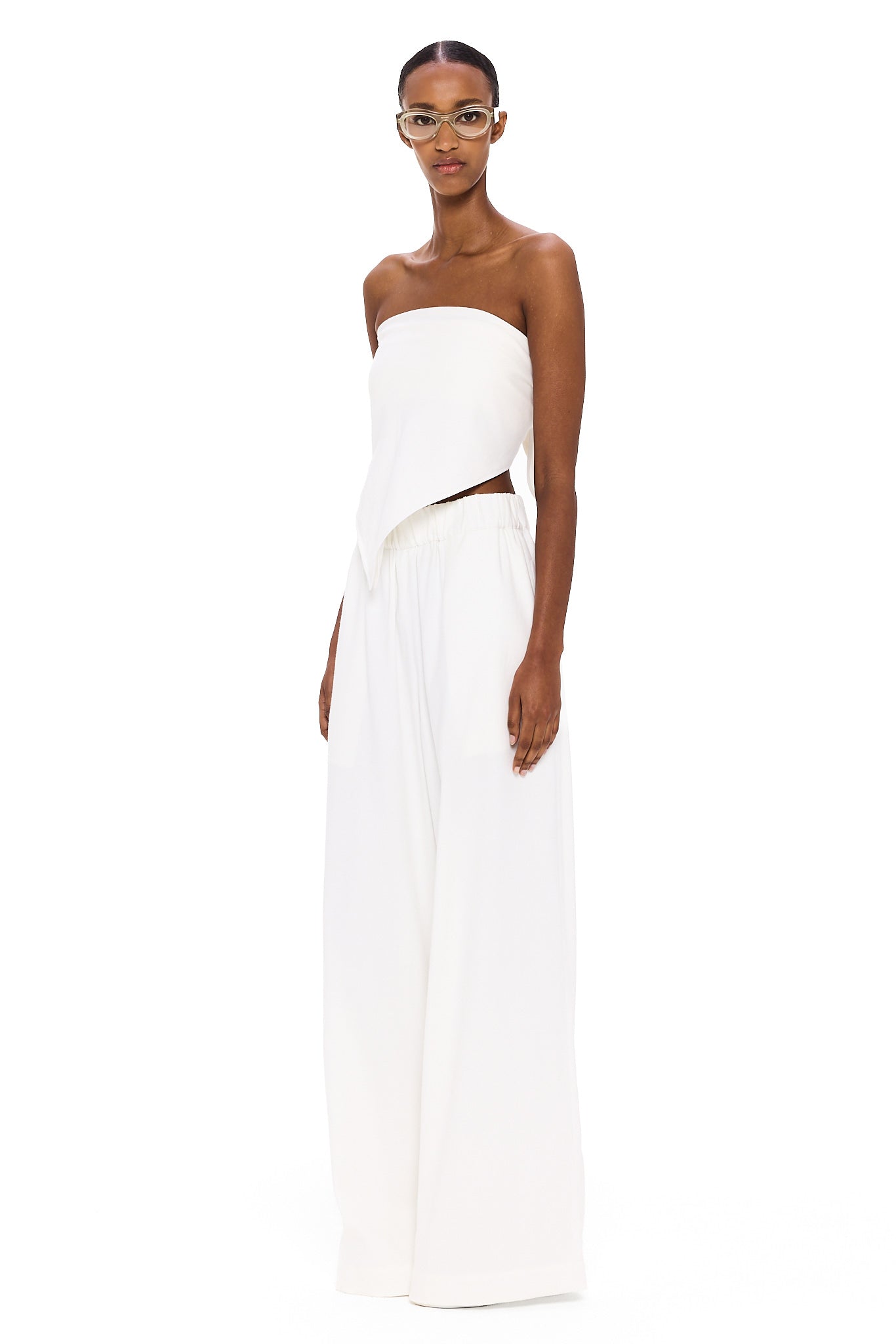 Dressed in The Triangle Top—a strapless white piece featuring an uneven hem—paired with wide-legged white pants, this individual stands against a white background, looking forward while wearing glasses; a modern showcase of sustainable fashion and the artistry of thoughtfully crafted, handmade clothing.