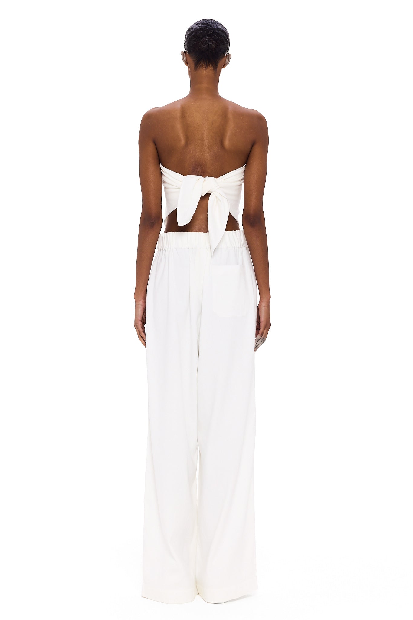 A person stands facing away, wearing The Triangle Top—a white strapless piece tied in a bow at the back—paired with wide-leg white pants featuring an elastic waistband. Handcrafted to embody sustainable fashion ideals, this minimalist ensemble is elevated by Moroccan weaving influences, celebrating the artistry of handmade clothing against a plain white background.