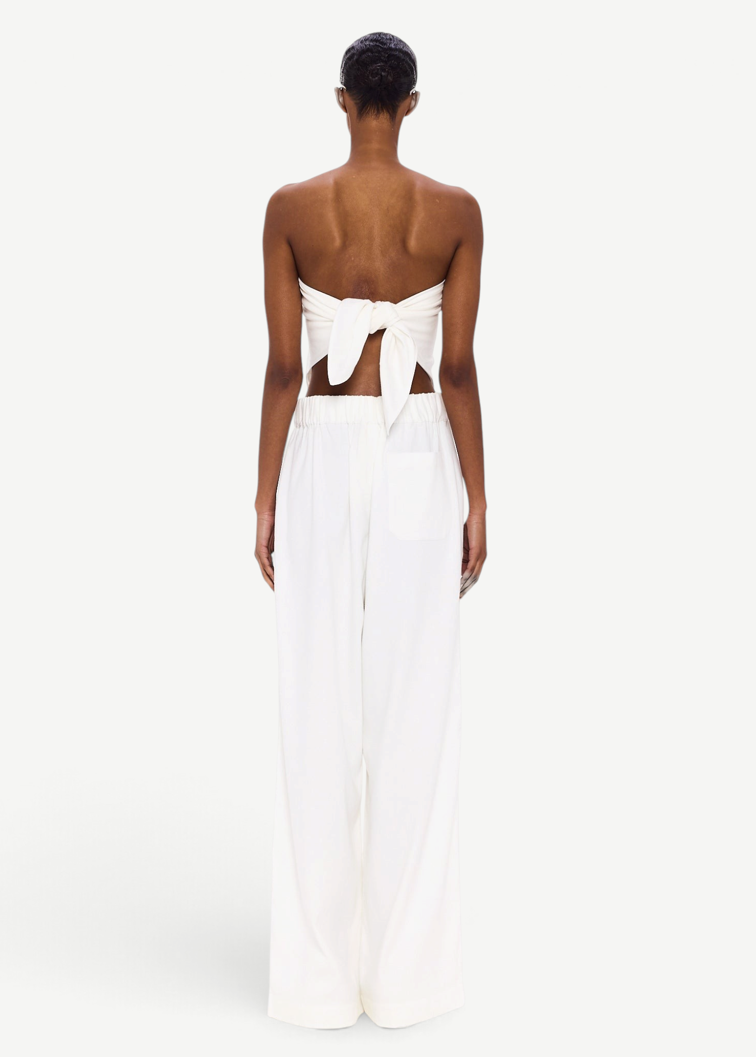 A person stands facing away, wearing The Triangle Top—a white strapless piece tied in a bow at the back—paired with wide-leg white pants featuring an elastic waistband. Handcrafted to embody sustainable fashion ideals, this minimalist ensemble is elevated by Moroccan weaving influences, celebrating the artistry of handmade clothing against a plain white background.