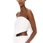 Wearing The Triangle Top—a white, asymmetrical strapless piece designed with sustainability in mind—and matching white pants, a woman with clear glasses stands against a plain white background, calmly gazing at the camera with one hand in her pocket.