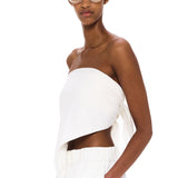 Wearing The Triangle Top—a white, asymmetrical strapless piece designed with sustainability in mind—and matching white pants, a woman with clear glasses stands against a plain white background, calmly gazing at the camera with one hand in her pocket.