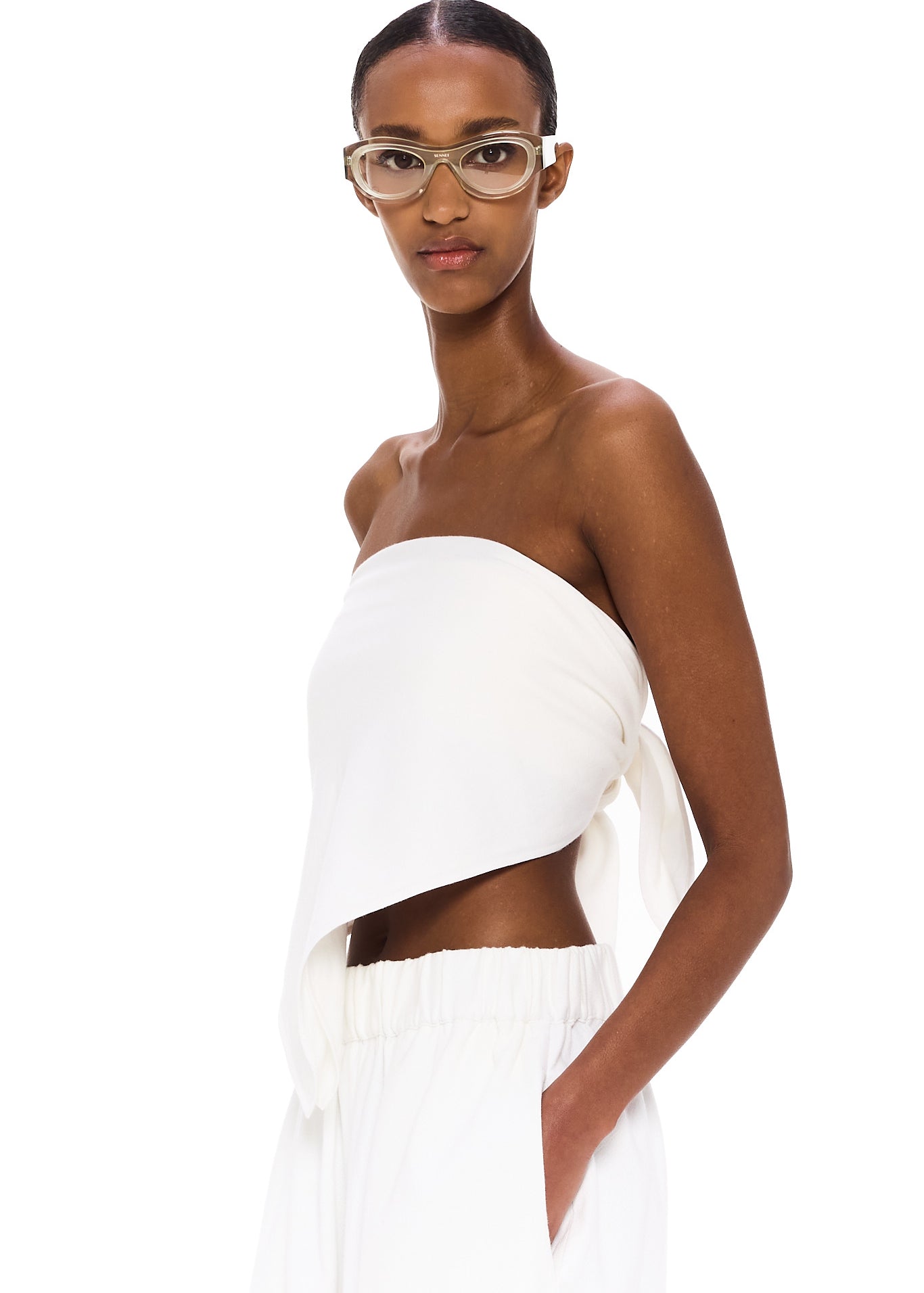 Wearing The Triangle Top—a white, asymmetrical strapless piece designed with sustainability in mind—and matching white pants, a woman with clear glasses stands against a plain white background, calmly gazing at the camera with one hand in her pocket.