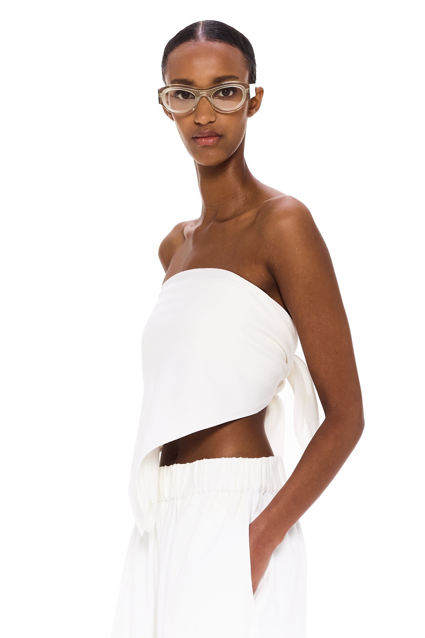Wearing The Triangle Top—a white, asymmetrical strapless piece designed with sustainability in mind—and matching white pants, a woman with clear glasses stands against a plain white background, calmly gazing at the camera with one hand in her pocket.