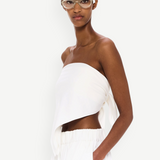 Wearing The Triangle Top—a white, asymmetrical strapless piece designed with sustainability in mind—and matching white pants, a woman with clear glasses stands against a plain white background, calmly gazing at the camera with one hand in her pocket.