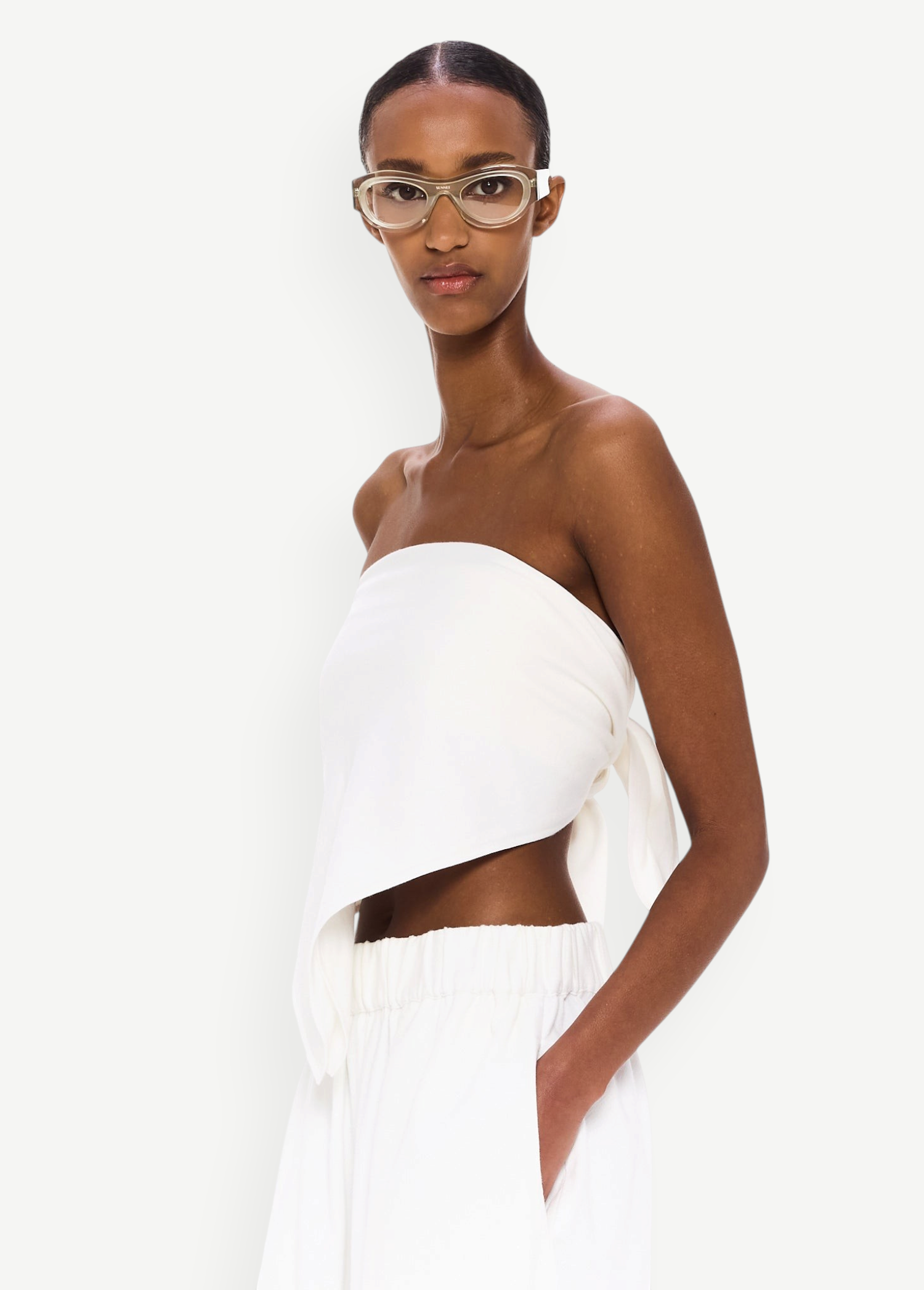 Wearing The Triangle Top—a white, asymmetrical strapless piece designed with sustainability in mind—and matching white pants, a woman with clear glasses stands against a plain white background, calmly gazing at the camera with one hand in her pocket.