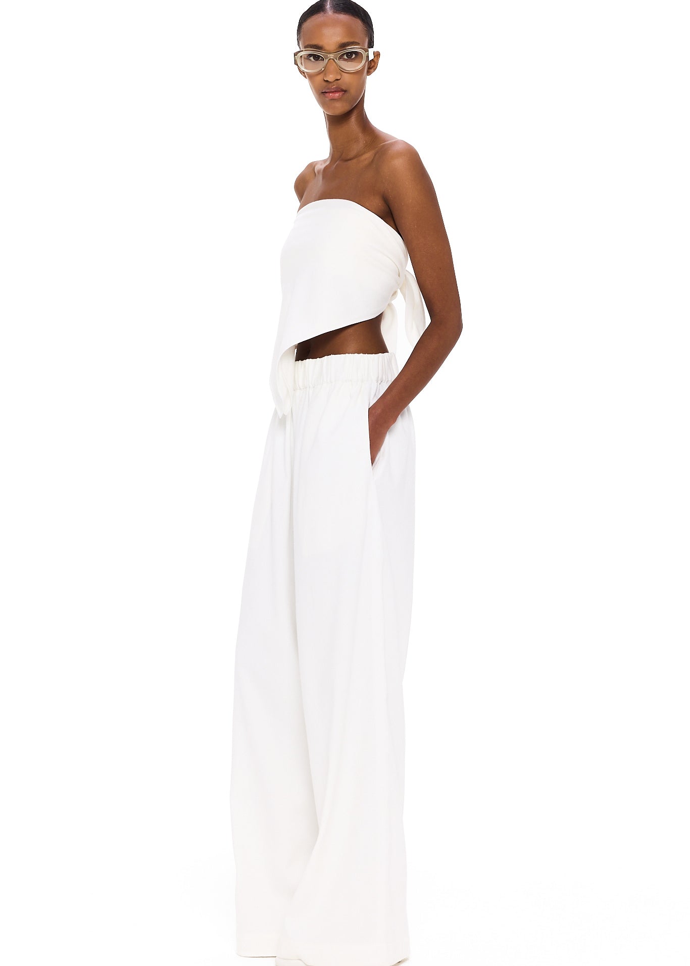 A person stands against a white background, wearing The Triangle Top—a white, strapless piece paired with loose white pants and clear glasses. With hands in their pockets and a confident gaze toward the camera, their outfit embodies sustainable fashion through handmade clothing inspired by Moroccan weaving traditions.