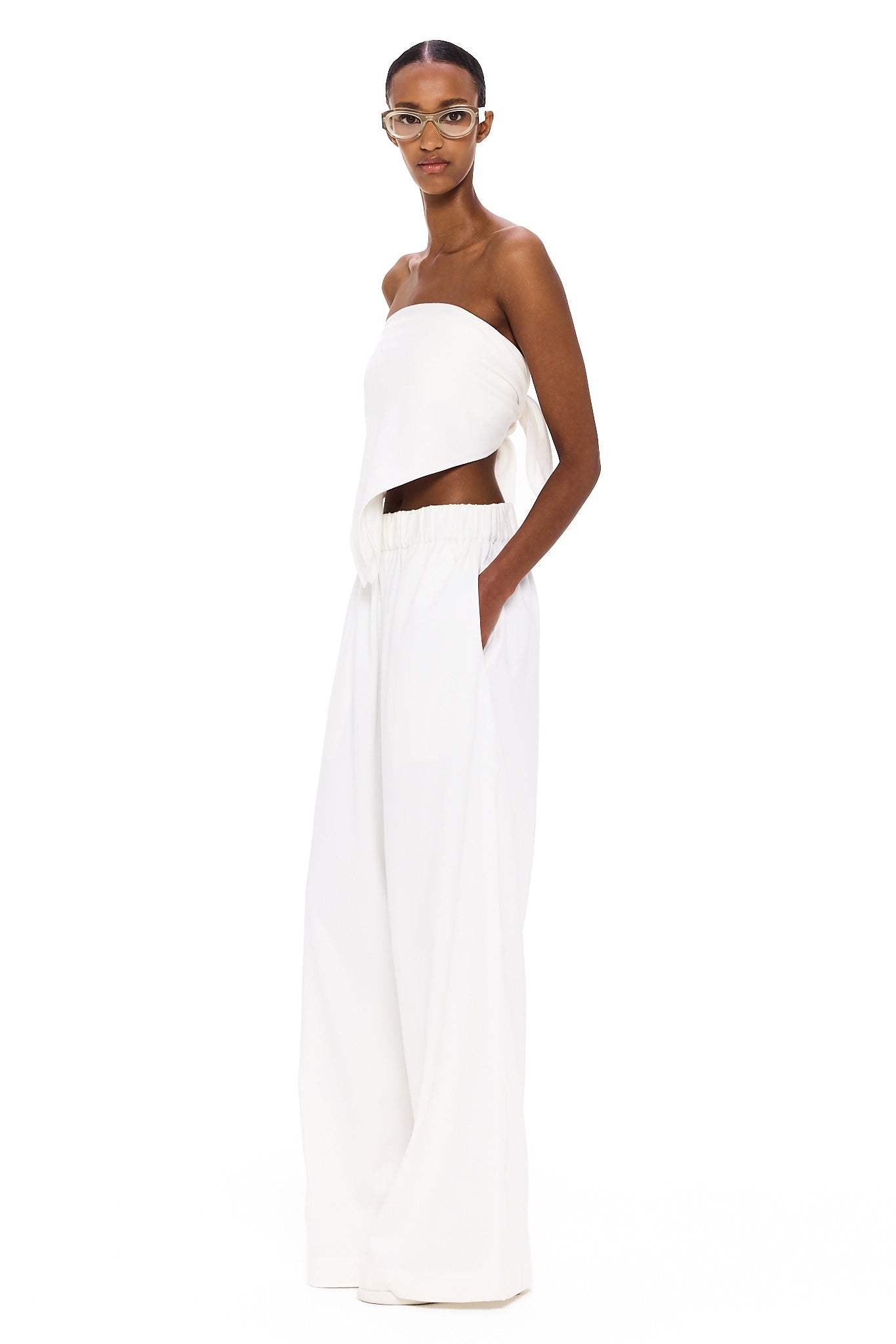 A person stands against a white background, wearing The Triangle Top—a white, strapless piece paired with loose white pants and clear glasses. With hands in their pockets and a confident gaze toward the camera, their outfit embodies sustainable fashion through handmade clothing inspired by Moroccan weaving traditions.