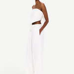 A person stands against a white background, wearing The Triangle Top—a white, strapless piece paired with loose white pants and clear glasses. With hands in their pockets and a confident gaze toward the camera, their outfit embodies sustainable fashion through handmade clothing inspired by Moroccan weaving traditions.