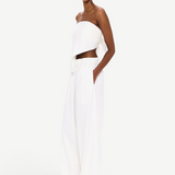 A person stands against a white background, wearing The Triangle Top—a white, strapless piece paired with loose white pants and clear glasses. With hands in their pockets and a confident gaze toward the camera, their outfit embodies sustainable fashion through handmade clothing inspired by Moroccan weaving traditions.