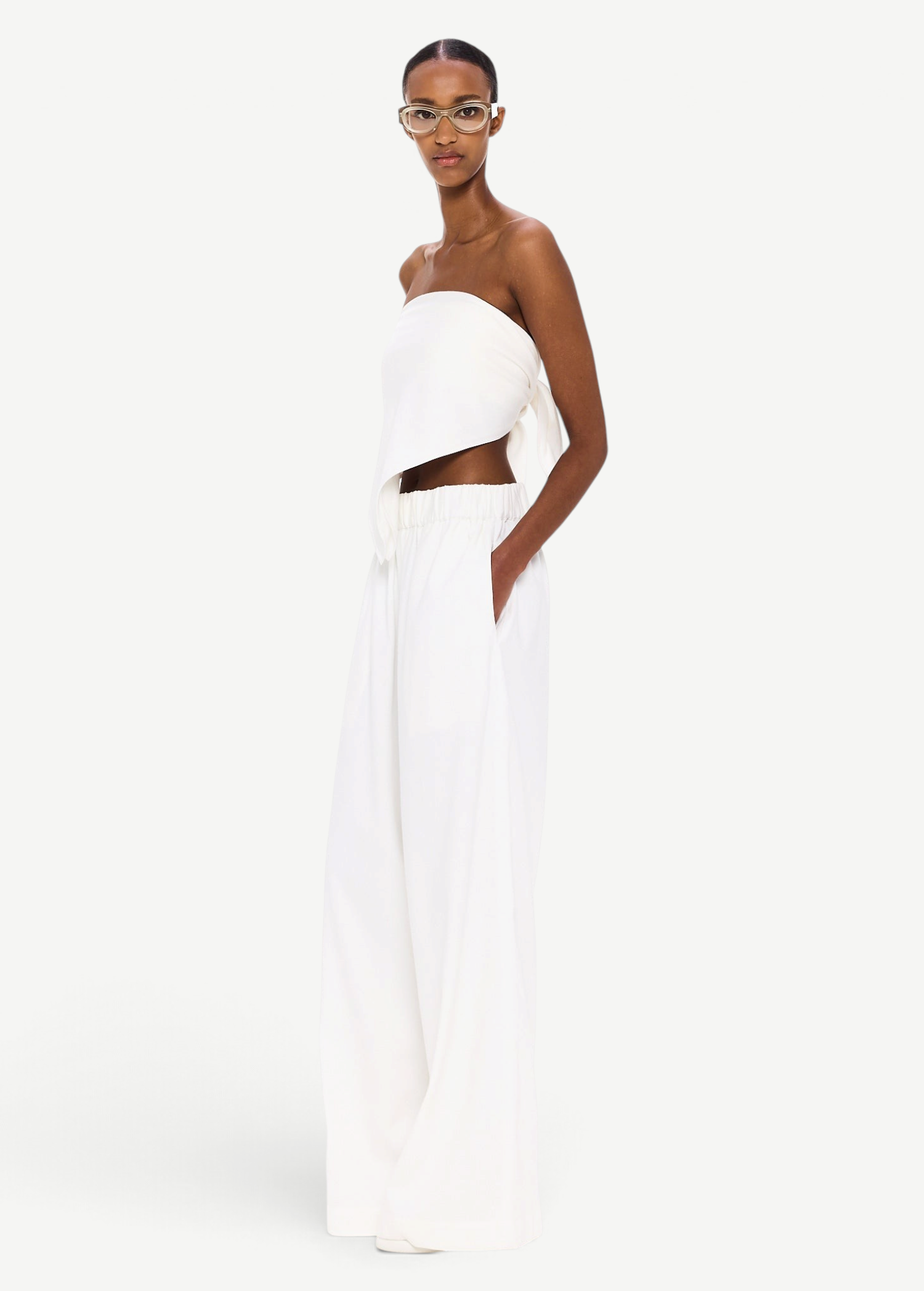 A person stands against a white background, wearing The Triangle Top—a white, strapless piece paired with loose white pants and clear glasses. With hands in their pockets and a confident gaze toward the camera, their outfit embodies sustainable fashion through handmade clothing inspired by Moroccan weaving traditions.
