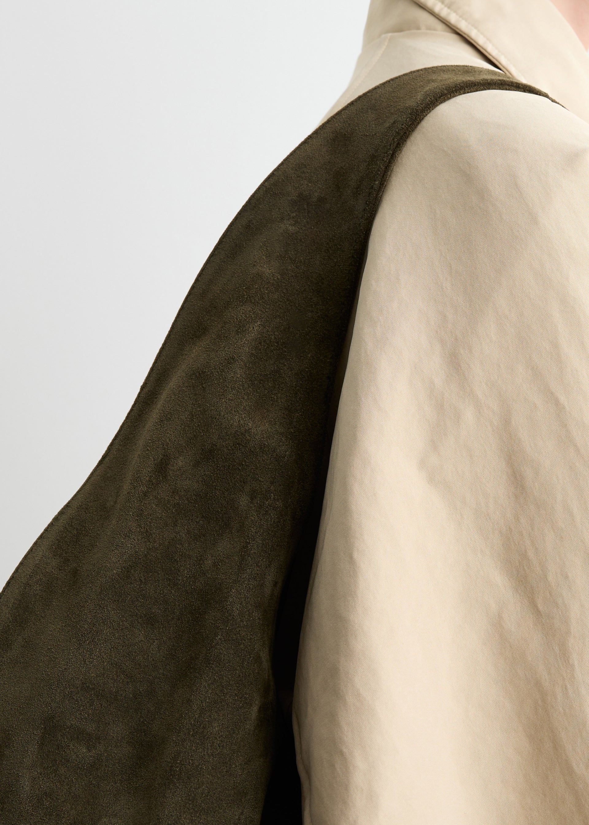 Close-up of a person wearing a beige trench coat with the Market Tote Bag, crafted from dark olive green suede, draped over their shoulder against a plain, light background.