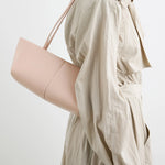 A person in a light beige trench coat carries the Boulevard Sling Bag, crafted from pale pink nappa leather with a minimalist design. The photo is cropped at the shoulders and mid-thigh against a neutral background.