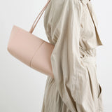 A person in a light beige trench coat carries the Boulevard Sling Bag, crafted from pale pink nappa leather with a minimalist design. The photo is cropped at the shoulders and mid-thigh against a neutral background.