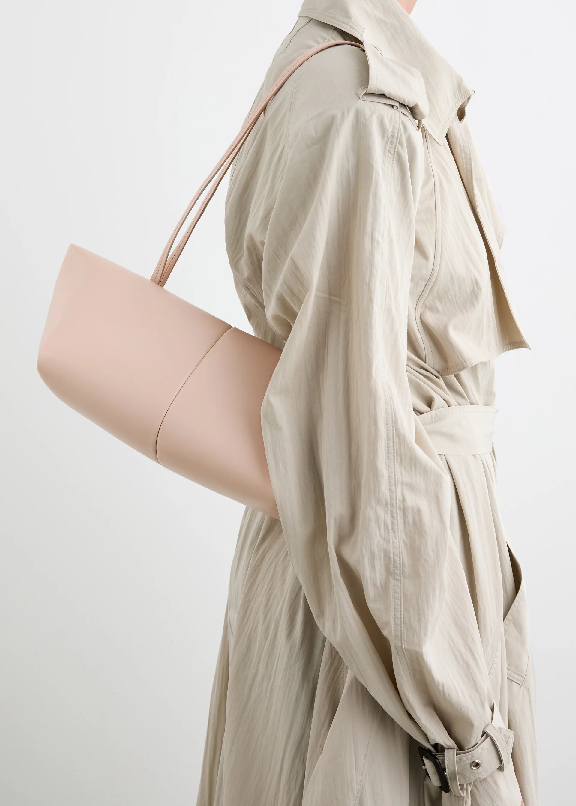 A person in a light beige trench coat carries the Boulevard Sling Bag, crafted from pale pink nappa leather with a minimalist design. The photo is cropped at the shoulders and mid-thigh against a neutral background.