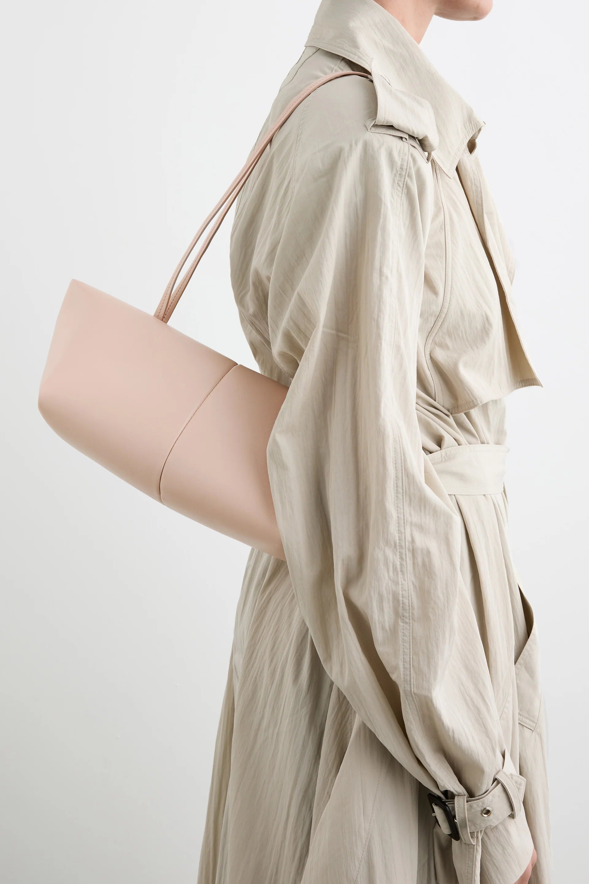 A person in a light beige trench coat carries the Boulevard Sling Bag, crafted from pale pink nappa leather with a minimalist design. The photo is cropped at the shoulders and mid-thigh against a neutral background.