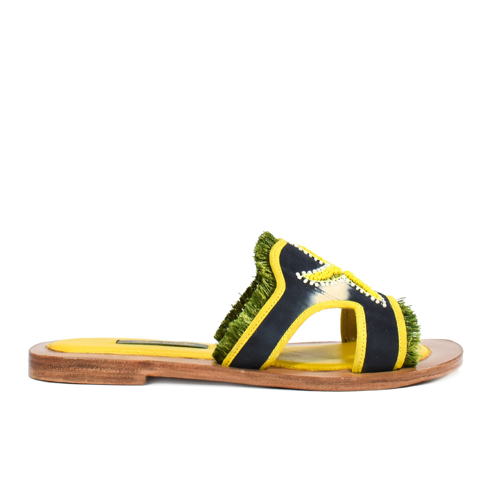 Introducing the Mazu Slide: a sustainable flat sandal featuring a wooden sole, yellow footbed, dark blue and yellow straps with green fringe trim, and accented by white stitching. Hand-embroidered leather detailing gives these slides a uniquely stylish finish.