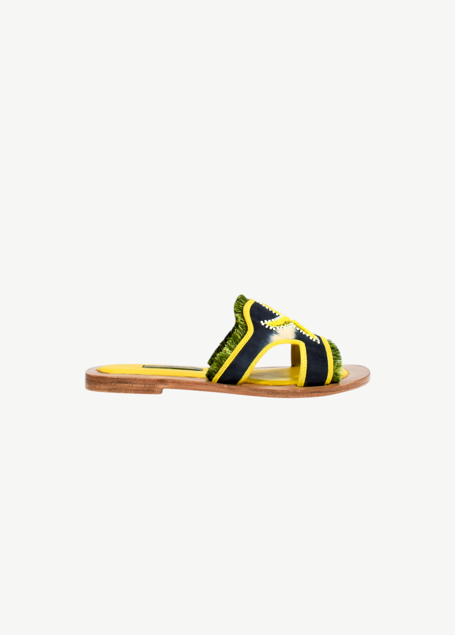 Introducing the Mazu Slide: a sustainable flat sandal featuring a wooden sole, yellow footbed, dark blue and yellow straps with green fringe trim, and accented by white stitching. Hand-embroidered leather detailing gives these slides a uniquely stylish finish.
