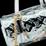 A silver Louis Vuitton handbag with embossed logos, adorned with the Heart Bag Charm—featuring a gold heart keychain, a colorful beaded accent, an elegant 14KT Gold Plate Chain, and a Martha Calvo Pendant—displayed against a black background.