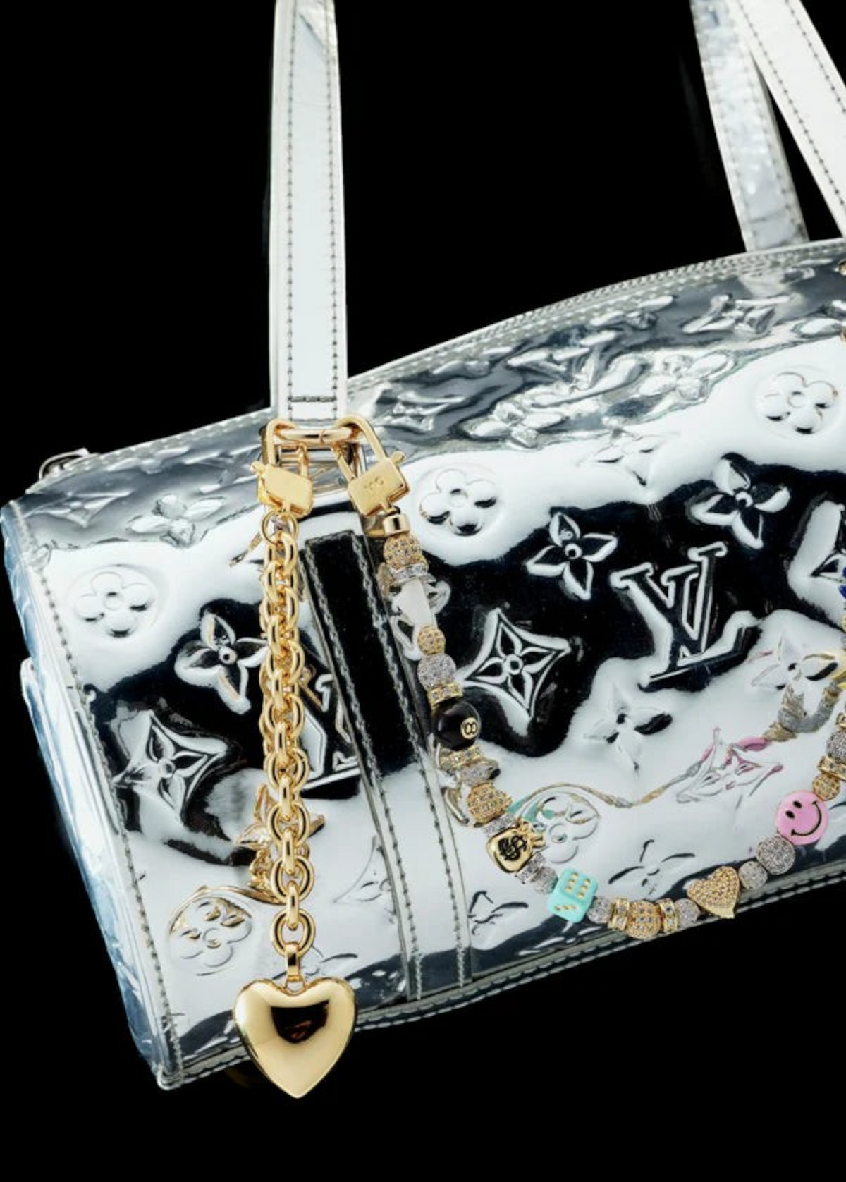 A silver Louis Vuitton handbag with embossed logos, adorned with the Heart Bag Charm—featuring a gold heart keychain, a colorful beaded accent, an elegant 14KT Gold Plate Chain, and a Martha Calvo Pendant—displayed against a black background.