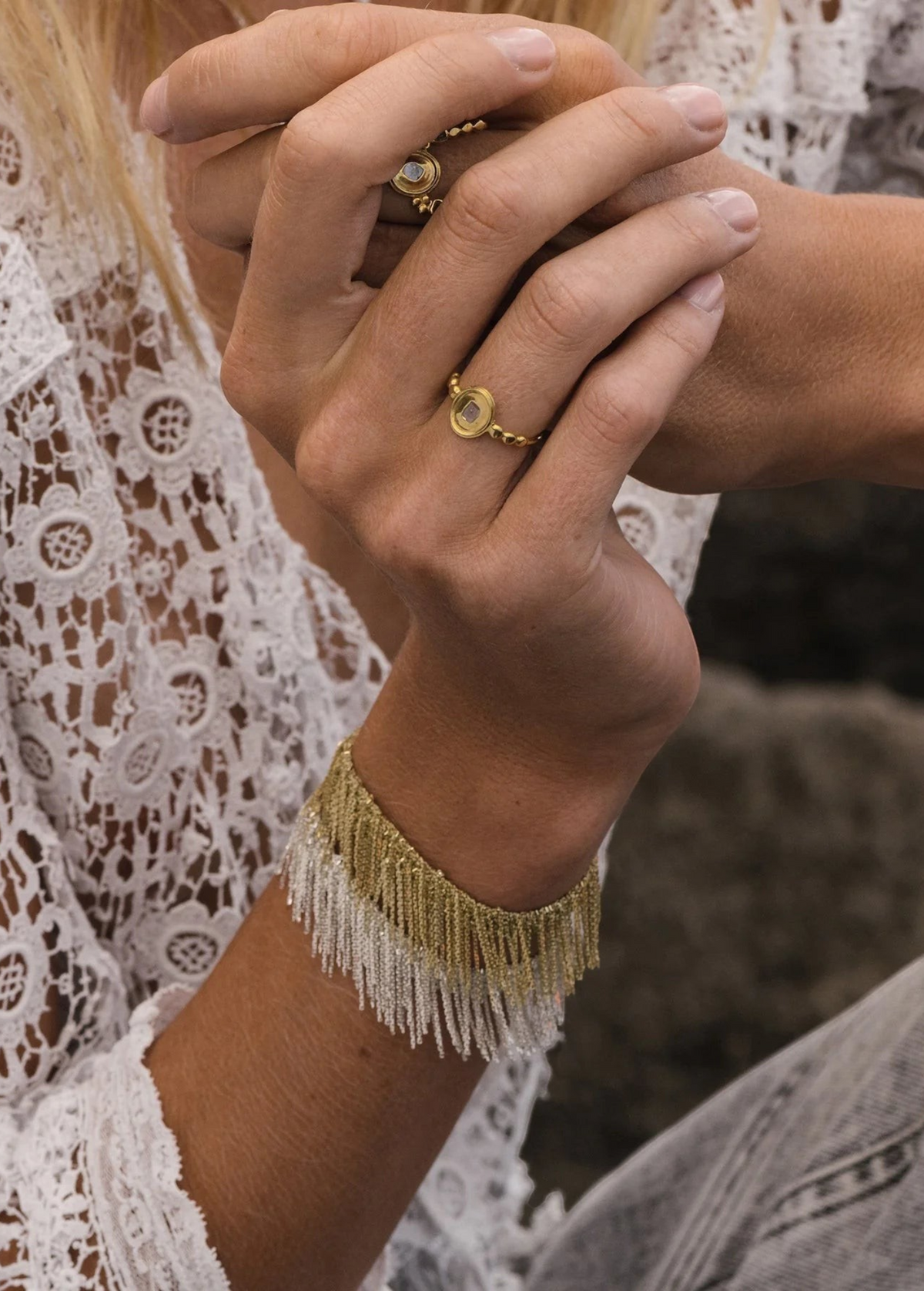 Close-up of a person’s hands wearing two gold rings with round stones and the N° 675 Silver Bracelet, dressed in a white lace top and jeans. Their nails are natural and short, adding a subtle touch perfect for everyday wear.
