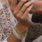 A person wearing lace clothing displays their hands adorned with two gold rings and the N° 675 Gold Bracelet, featuring a delicate gold finish and white beaded fringe. The background is blurred, emphasizing the jewelry and intricate lace details.