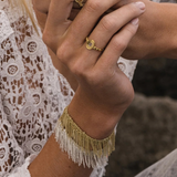 A person wearing lace clothing displays their hands adorned with two gold rings and the N° 675 Gold Bracelet, featuring a delicate gold finish and white beaded fringe. The background is blurred, emphasizing the jewelry and intricate lace details.