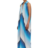 A person stands against a plain white background, wearing The Halter Dress Small Point Collar—a sleeveless, flowing halter dress featuring bold diagonal blue, white, and gray stripes. Crafted from handwoven cotton that highlights Moroccan craftsmanship, they have styled hair and dramatic eye makeup.