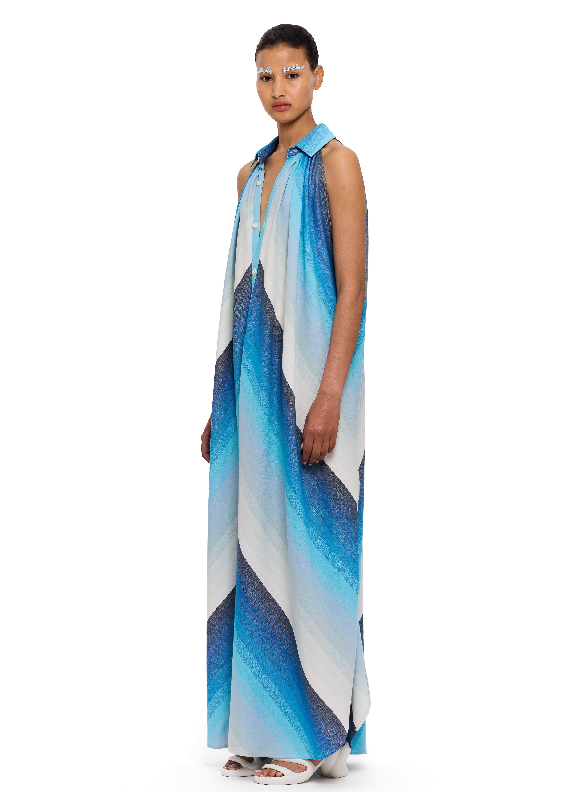 A person stands against a plain white background, wearing The Halter Dress Small Point Collar—a sleeveless, flowing halter dress featuring bold diagonal blue, white, and gray stripes. Crafted from handwoven cotton that highlights Moroccan craftsmanship, they have styled hair and dramatic eye makeup.