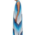 A woman stands in profile against a white background, wearing The Halter Dress Small Point Collar—a long, sleeveless dress featuring blue and white chevron stripes. She pairs the look with white shoes and slicked-back hair accented by blue eye makeup. The handwoven cotton fabric reflects exquisite Moroccan craftsmanship.