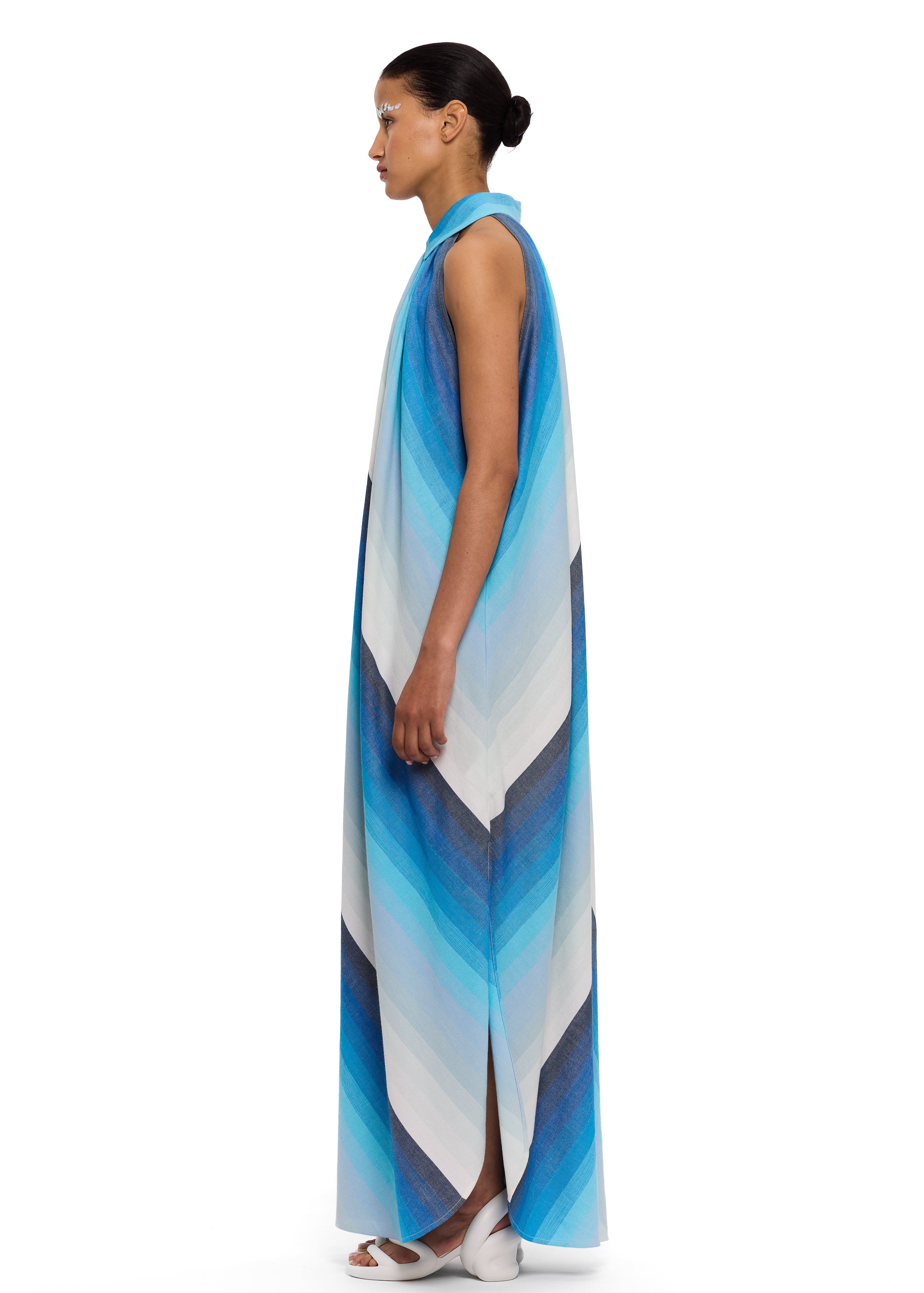 A woman stands in profile against a white background, wearing The Halter Dress Small Point Collar—a long, sleeveless dress featuring blue and white chevron stripes. She pairs the look with white shoes and slicked-back hair accented by blue eye makeup. The handwoven cotton fabric reflects exquisite Moroccan craftsmanship.