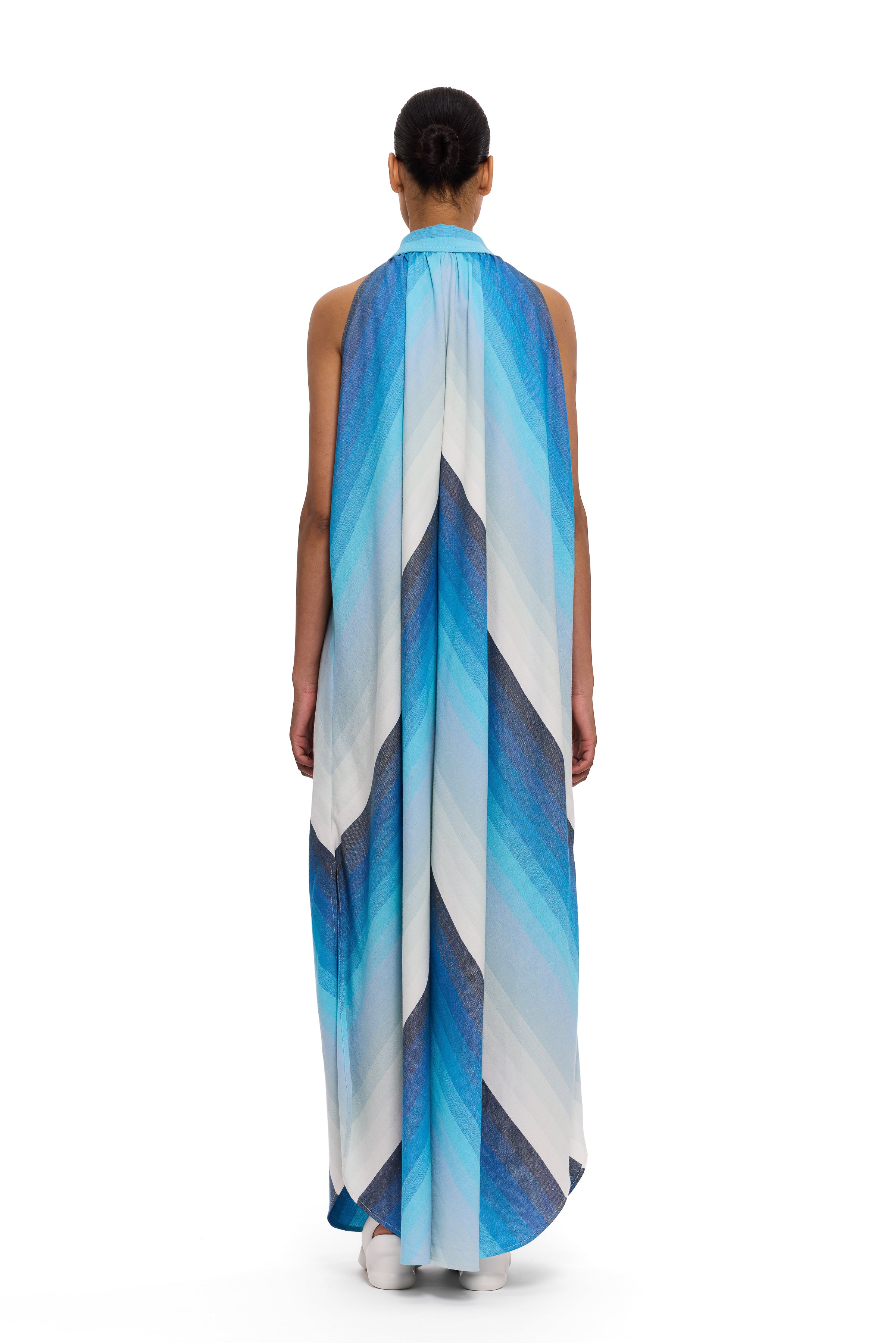 A person stands facing away, wearing The Halter Dress Small Point Collar—a long, flowing halter dress featuring blue and white diagonal stripes and crafted from handwoven cotton. Their hair is tied back in a neat bun, set against a plain white background.