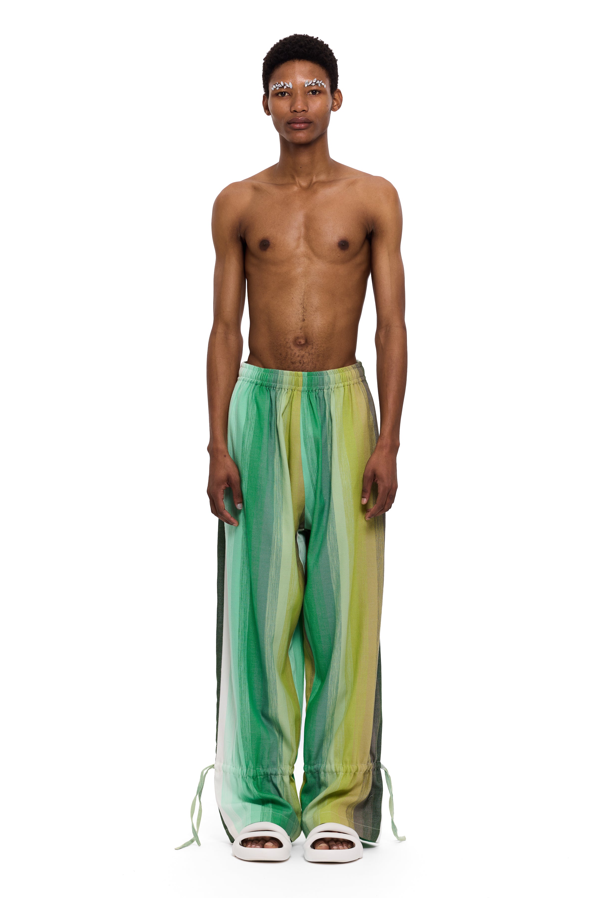 A shirtless person wears The Parachute Pant—green gradient wide-leg parachute pants made from handwoven cotton—paired with white sandals and adorned with pearls or beads above their eyebrows, standing facing forward against a plain white background and capturing the timeless aesthetic of Moroccan craftsmanship.