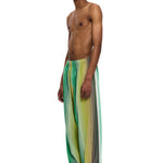 A shirtless person stands against a white background, confidently showcasing The Parachute Pant—loose, sheer green and yellow striped pants made from handwoven cotton—paired with white sandals and decorative face makeup, highlighting exceptional Moroccan craftsmanship.