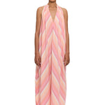 A person stands wearing The Halter Dress V-Neck, a loose, sleeveless halter dress featuring pink, peach, and cream diagonal stripes crafted from handwoven cotton, paired with matching sandals. The background is plain white.