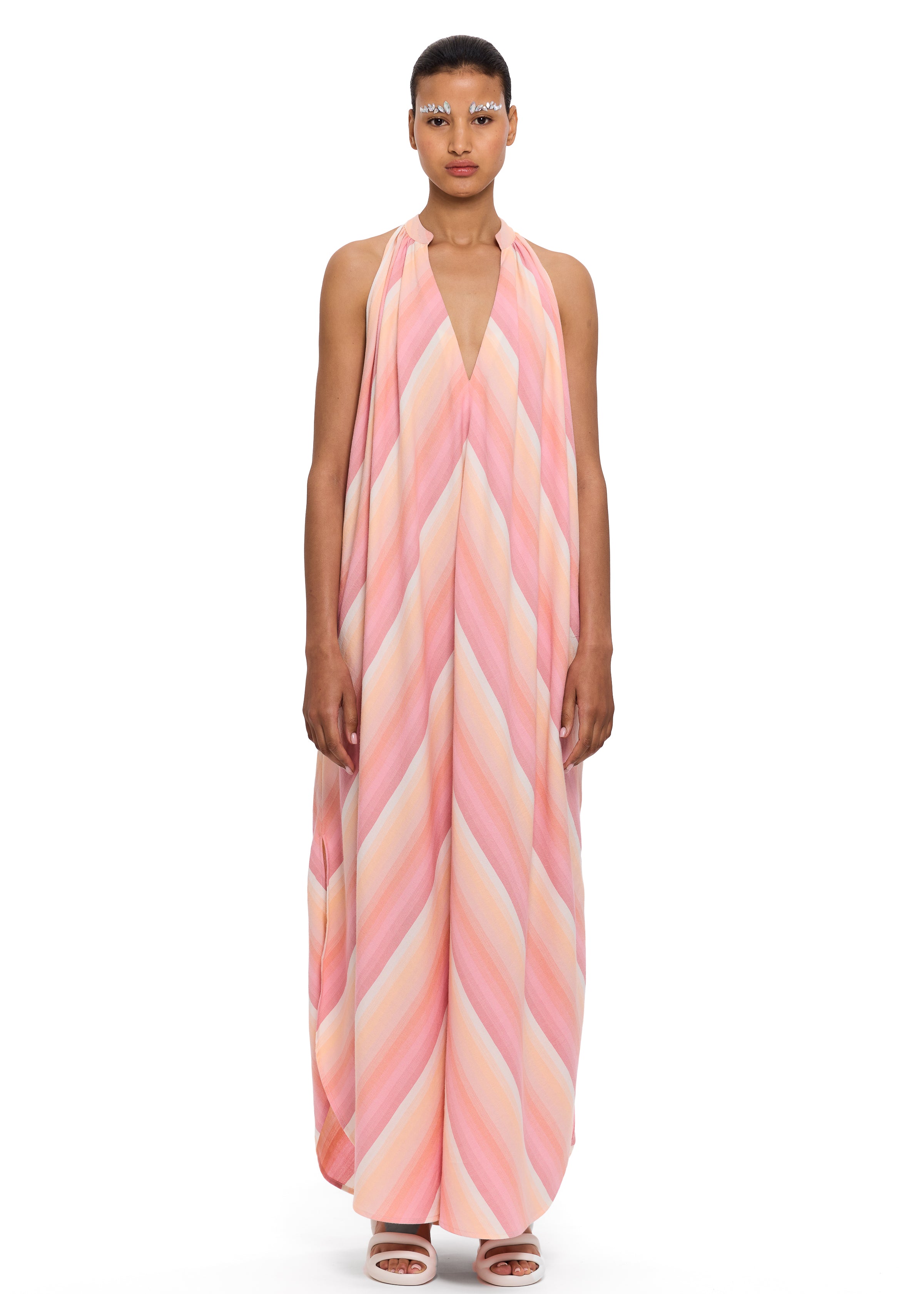 A person stands wearing The Halter Dress V-Neck, a loose, sleeveless halter dress featuring pink, peach, and cream diagonal stripes crafted from handwoven cotton, paired with matching sandals. The background is plain white.