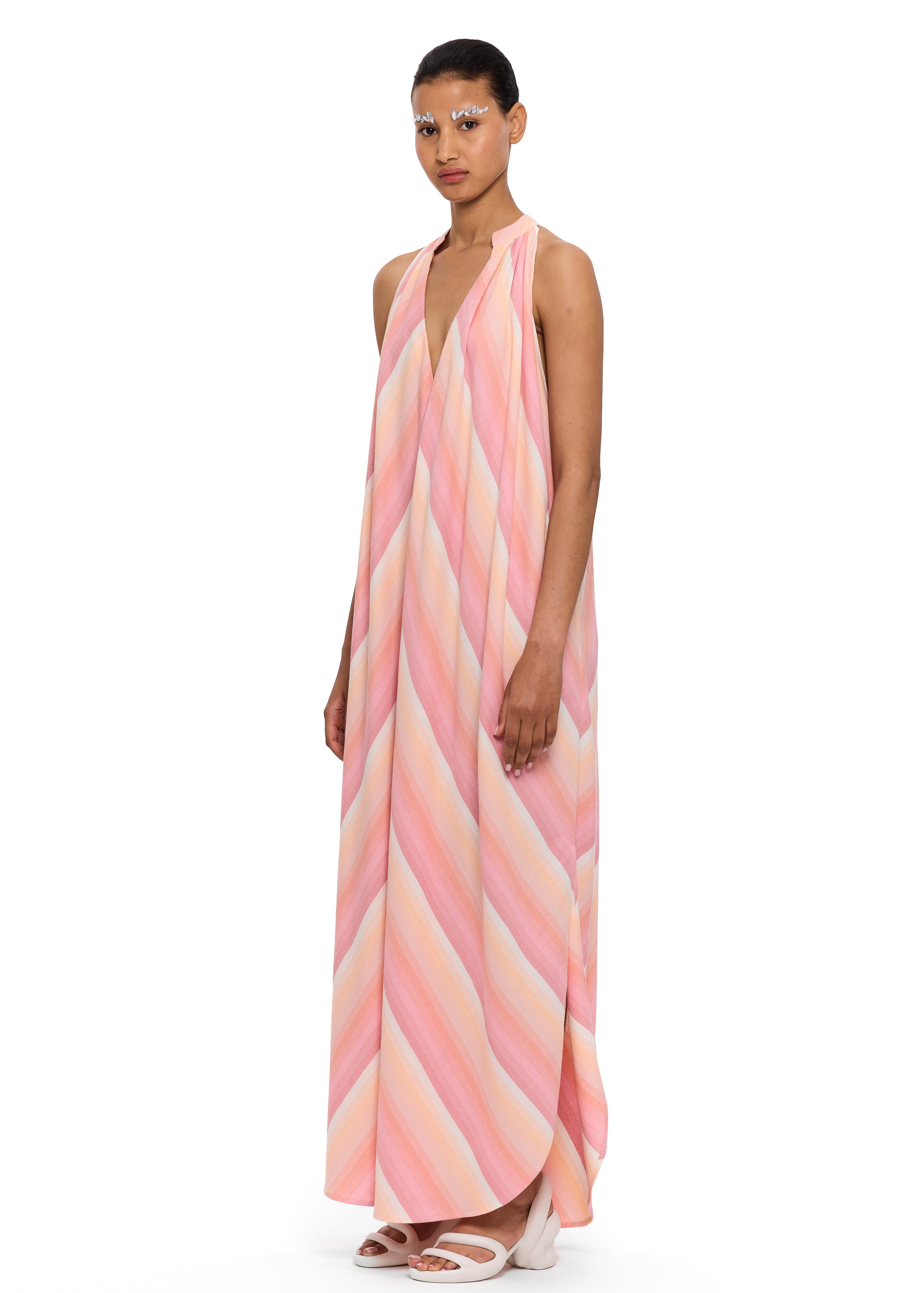 A person stands against a white background, wearing The Halter Dress V-Neck—a loose, flowy, sleeveless dress with pink and peach stripes crafted from handwoven cotton—paired with white shoes and distinctive white eye makeup.
