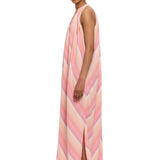 A person wearing The Halter Dress V-Neck—a long, sleeveless dress with pastel stripes, side slits, and a halter neckline—stands sideways against a white background. Their hair is pulled back and they wear decorative makeup above their eyes, evoking the relaxed charm of Marrakech. White sandals complete the look.