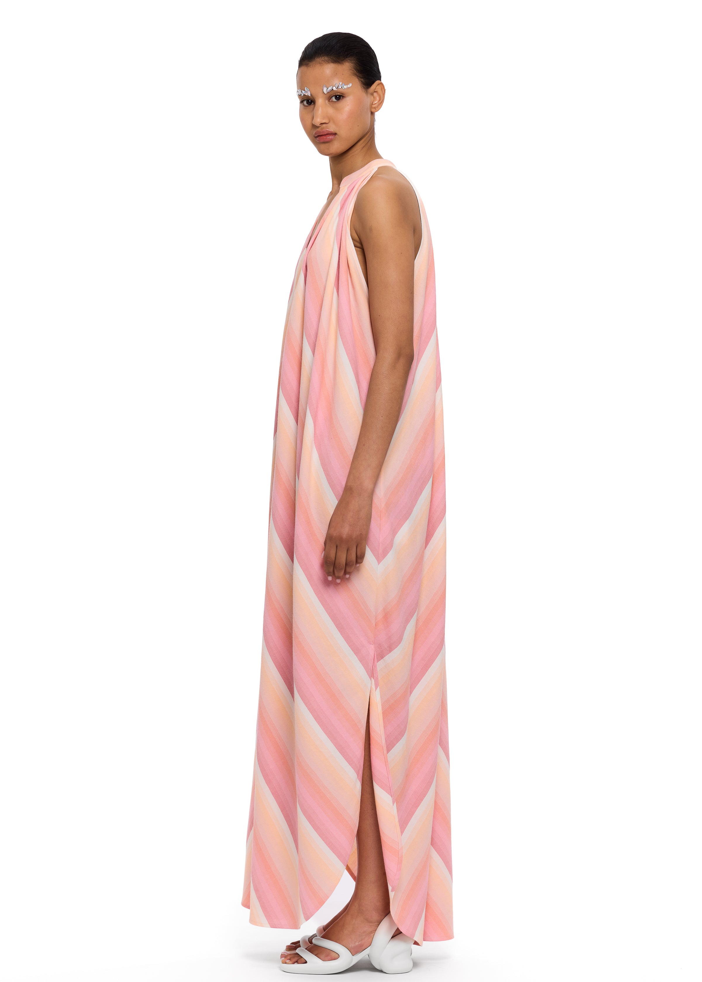 A person wearing The Halter Dress V-Neck—a long, sleeveless dress with pastel stripes, side slits, and a halter neckline—stands sideways against a white background. Their hair is pulled back and they wear decorative makeup above their eyes, evoking the relaxed charm of Marrakech. White sandals complete the look.