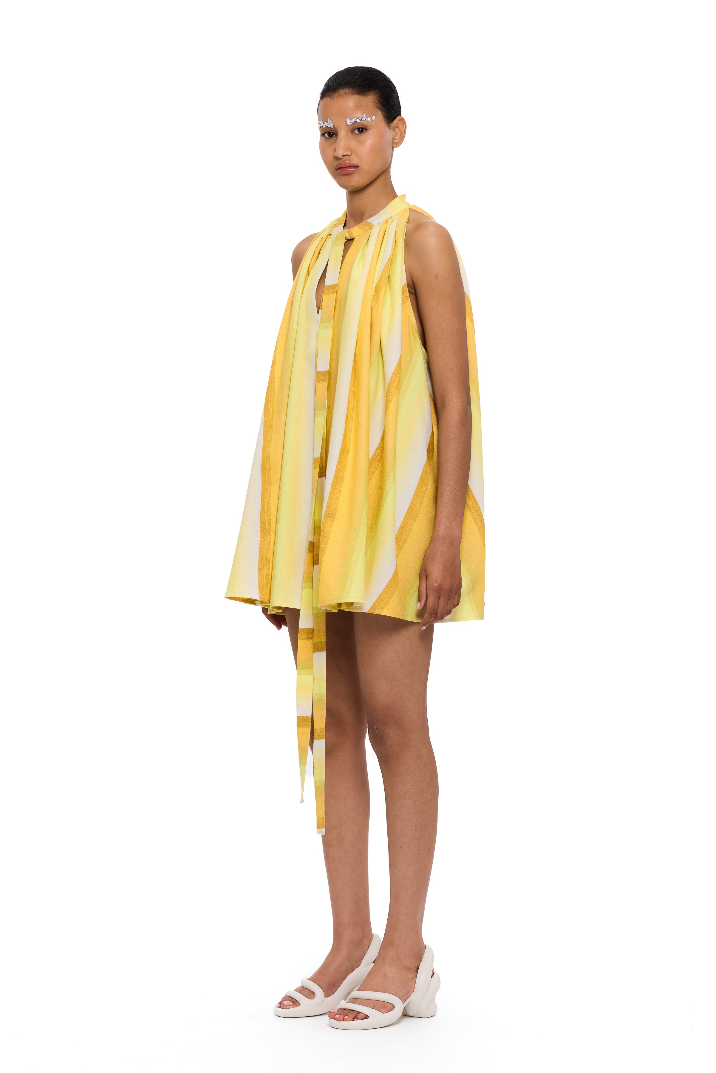 A person wearing The Mini Cape Dress—a yellow and white striped, sleeveless, loose-fitting handwoven cotton dress featuring a necktie detail—paired with white shoes and decorative eye makeup stands against a plain white background, embodying the essence of sustainable fashion.